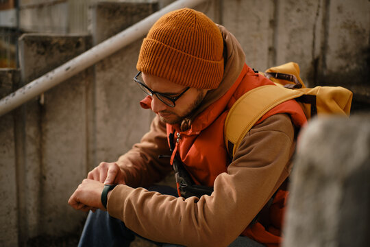 Close-up of man in orange beanie and vest checking time on smartwatch while sitting on stairs
