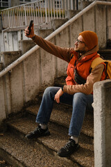 Fototapeta premium Man sitting on stairs taking selfie with smartphone, wearing orange vest and beanie. Travel blogger concept