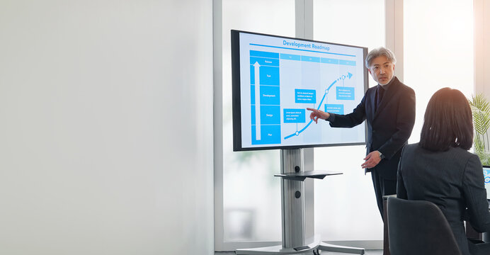 A middle-aged male business person giving an explanation in front of a large screen monitor
