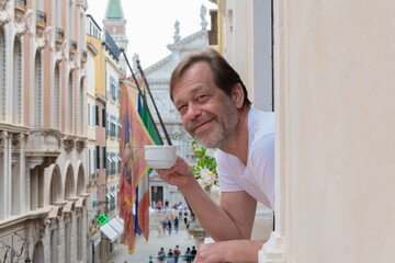 A man with a cup of coffee stands at the window looking out from the balcony onto the streets of Venice's historic centre.