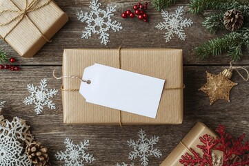Gift box with a blank tag and space for writing. Seasonal winter holidays theme. Getting ready for the festive season. Overhead view of holiday ornaments on a rustic wooden surface.