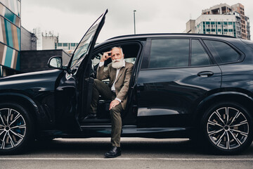 Stylish senior businessman confidently posing on the city streets next to a luxury black car during a bright day