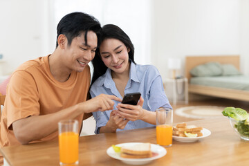 Asian couple smiling and using smartphone together during breakfast at home. Concept of modern love, digital lifestyle, joyful relationship, and casual morning moment in cozy setting.