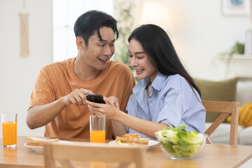 Asian couple laughing and enjoying time together while using smartphone during breakfast at home. Concept of happiness, digital lifestyle, relationship bonding, and morning routine.