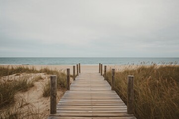 Fototapeta premium A worn wooden walkway descends to a peaceful sandy shore with gentle waves and an overcast sky.
