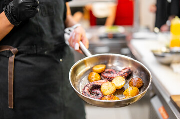 Chef holding pan with roasted octopus tentacles and baby potatoes in professional kitchen. Culinary presentation, seafood dish, restaurant cooking concept.
