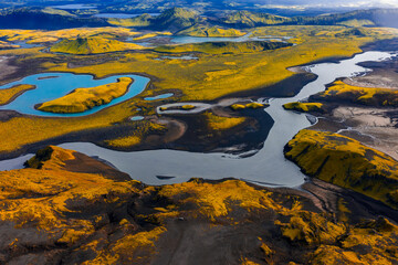 Aerial view of Iceland's yellow moss covered terrain, dark volcanic soil, winding river, blue lake with an island, craters, and distant mountains.