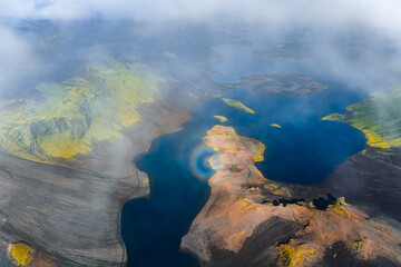 Aerial view of a blue lake in Iceland surrounded by volcanic terrain, green moss, craters, and a circular rainbow under misty clouds.