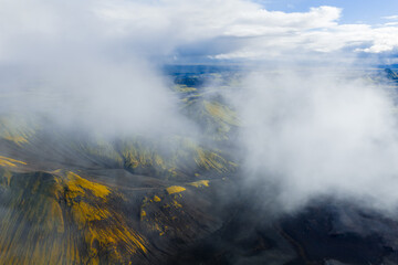 Aerial view of Iceland's volcanic landscape with dark soil, vibrant moss, craters, and ridges, partially veiled by low hanging clouds.