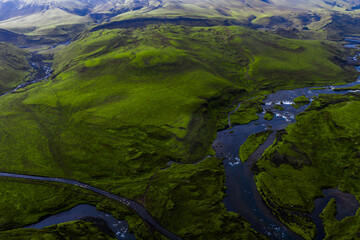 Aerial perspective of Iceland's lush green hills, winding rivers, and a road cutting through the terrain, with mountains and waterfalls in the distance.