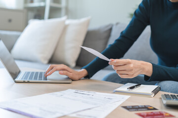 Stressed asian young business woman, employee using laptop computer to calculate expenses, hand holding bills and receipt for to payment on table at home. Financial, finance of banking concept.