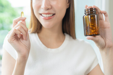 Fototapeta premium Dietary supplement concept, happy asian young woman hand holding fish oil pills, drugs medical tablet pouring capsules from medication bottle, take vitamin treatment for skin, hair at home, healthcare