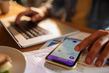 Young man using a banking app on his smart phone while working from home on his laptop in the kitchen