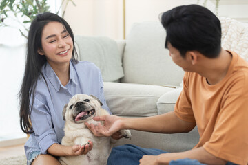 Happy Asian couple playing with adorable pet dog in cozy living room. Concept of love, relationship bonding, family lifestyle, pet companionship and joyful moments at modern home.