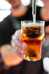 Close-up of a hand holding a glass of amber beer being poured from a tap, with foam forming on top. Frosted glass, blurred background. Concept of draft beer, bar service, refreshment.