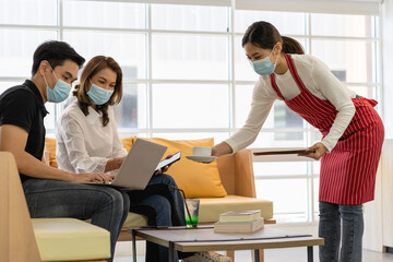 A man in black polo shirt and a woman in white shirt sitting on a couch in the coffee shop. A woman in a red apron serves a cup of coffee to a woman in a white shirt. A man using a laptop.