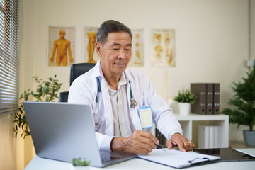 Experienced male doctor n white coat filling out patient records in a bright clinic with anatomical charts.
