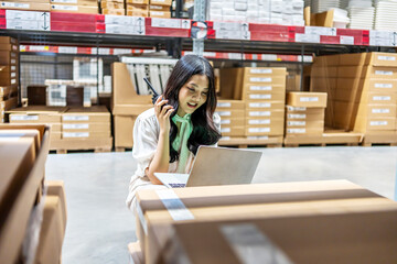 lively warehouse, dedicated young asian woman meticulously checks inventory details on her laptop while engaged in a conversation for radio communication, ensuring smooth operations and accuracy