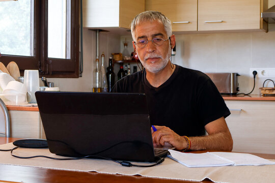 Casual middle-aged man working remotely on laptop at kitchen table, home office lifestyle. - Powered by Adobe