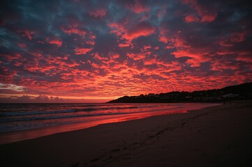 Obraz premium Vibrant crimson sunset over the ocean with a cloudy sky and crashing waves, small coastal homes in the distance, evoking a sense of adventure and picturesque views.