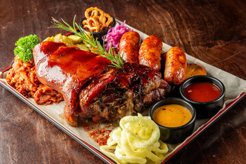 Rustic meat platter with glazed pork knuckle, grilled sausages, roasted potatoes, sauerkraut, pickles, sauces, and pretzels on parchment paper. Traditional hearty restaurant meal.