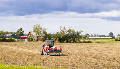 Obraz premium Agricultural tractor plowing a field under a cloudy sky, with green grass in the foreground and distant farm buildings creating a serene rural landscape