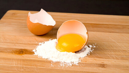 A cracked egg with a bright yellow yolk sits on a pile of white flour on a wooden cutting board.