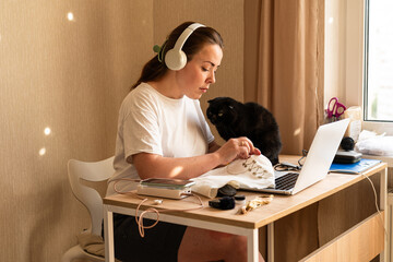 Woman Embroidering While Wearing Headphones at Home Desk. Senior black cat sits beside her, while a laptop rests on the desk
