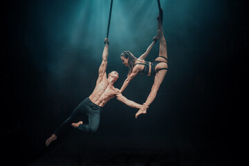Male and female aerialists perform on straps mid-air, the woman holding a vertical split while gripping the strap, supported by the man&rsquo;s arm under dramatic blue stage lighting.