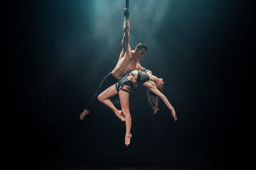 Male and female aerialists performing an elegant duo pose on straps under dramatic blue spotlights....
