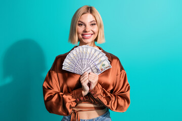 Obraz premium Elegant woman holding fan of cash bills in hand on turquoise background wearing stylish blouse displaying excitement