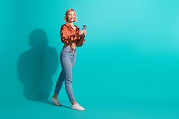Fototapeta premium Charming young woman in casual outfit using smartphone against turquoise backdrop promoting a stylish and happy lifestyle