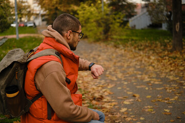 Man with backpack checking smartwatch while sitting on park bench with fallen leaves