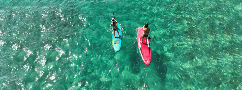 Aerial drone ultra wide photo of couple practice Stand Up Paddle board or SUP surf in tropical exotic island bay with emerald crystal clear sea - Powered by Adobe