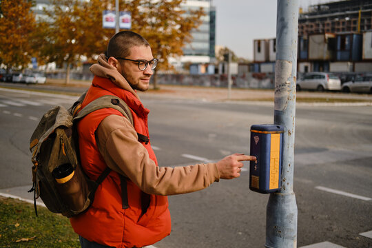 Man with backpack presses pedestrian crossing button on city street in Belgrade. Modern buildings and cars in background