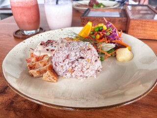 A plate with mixed grain rice, sliced grilled chicken with sauce, and a colorful vegetable salad garnished with an orange slice, with drinks in the background.