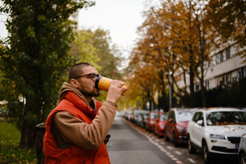 Young man in glasses drinking hot tea or coffee from yellow thermos on a city street with autumn...