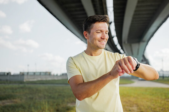 Man checks watch while enjoying time outdoors under a large bridge in a green space nearby urban area - Powered by Adobe