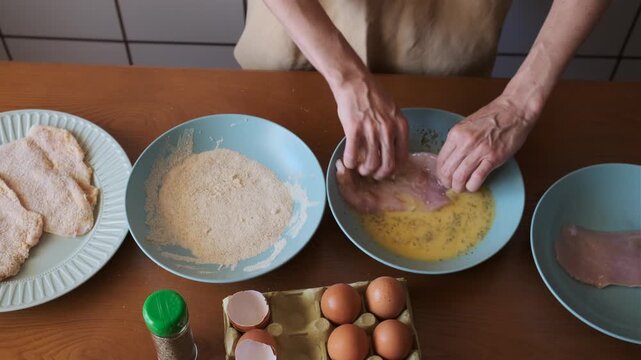 Overhead closeup of an adult woman dipping chicken fillet in egg wash and pressing it into breadcrumbs at a home kitchen counter.
