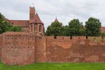 Historic brick wall and medieval buildings in Malbork Castle, Poland, surrounded by lush greenery under a cloudy sky