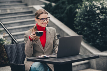 Young professional businesswoman with coffee cup working outdoors on laptop at stylish urban cafe