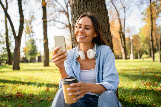 Woman looking at her mobile phone while holding a coffee cup. Female student enjoying beverage in park while taking a selfie using smartphone online app, browsing internet, listening music