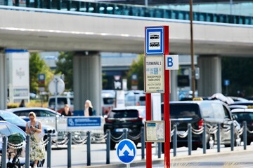 Fototapeta premium Modern airport terminal with road signs, parking directions, passengers with luggage, shuttle bus stops, and glass architecture highlighting international travel and transportation.