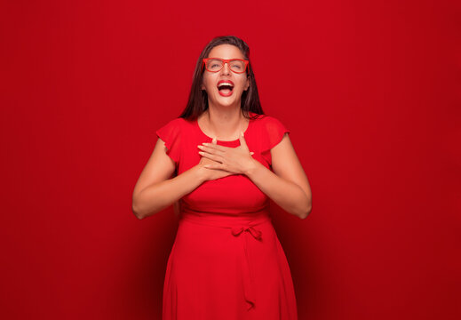 Thank you. Grateful young woman looking up with hand on his chest, isolated on red background. Rejoicing girl celebrates success.