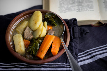 Homemade beef and vegetable stew with potatoes, carrots, and kale served in a rustic bowl beside an open cookbook — comforting winter meal with hearty ingredients and warm, traditional food styling