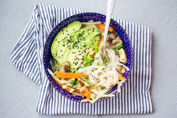 Vibrant noodle bowl with avocado, mushrooms, carrots, and cabbage topped with black sesame seeds...