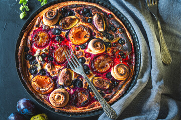A tart or open pie with blueberries, figs, prunes and apricots in a baking dish on a gray background with a fork and napkin on the table. Top view. High quality photo