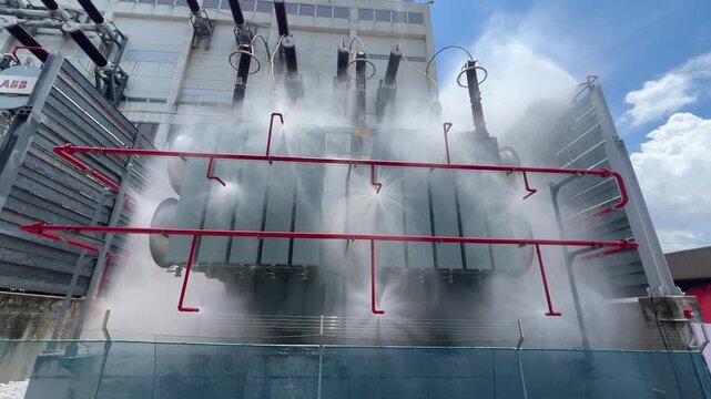 Industrial transformer substation fire protection system water spray mist drill, heavy mist cools metal structure, safety test with red pipe network and dramatic steam cloud outdoors bright sky