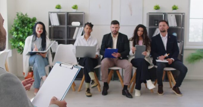Male team leader or business coach with clipboard talking with colleagues sitting in row during team meeting. Company employees taking notes while listening to speaker during presentation or training.