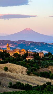 A picturesque Tuscan landscape at sunset, featuring rolling hills, a village nestled within the valley, and a majestic volcano in the background bathed in warm hues.